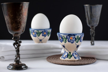 Still life of two white eggs in egg stands with white napkins near it and two sterling silver wine...