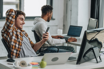 cheerful programmer using smartphone while sitting at workplace near african american colleague