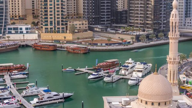 Yachts in Dubai Marina flanked by the Al Rahim Mosque and residential towers and skyscrapers aerial timelapse. Modern high rise skyline with boats and wooden dhows