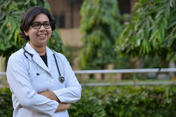 Portrait of a confident young female professional doctor wearing white coat and stethoscope in an outside setting in a park or hospital lawns in New Delhi, India