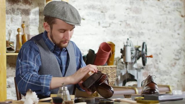 Male shoemaker in flat cap and vest polishing leather boot with brush at work desk in his studio and then looking at camera