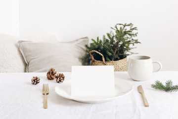 Festive Christmas, winter table setting. Golden cutlery, spruce tree branch, linen tablecloth, porcelain dinner plate, cup of coffee and silk pine cones on white table background. Blank card mockup.