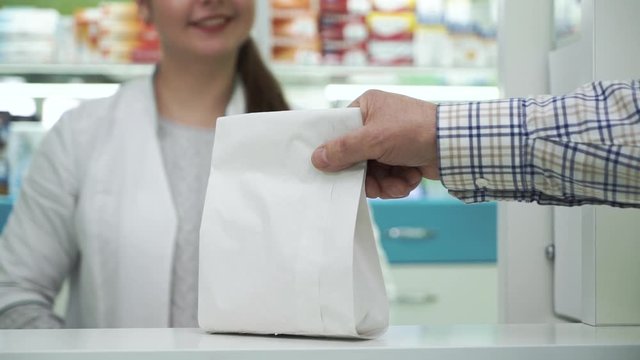 Smiling pharmacist giving the white paper bag with medications to the male customer. Purchase process in the pharmacy. Healthcare and traditional medicine concept.