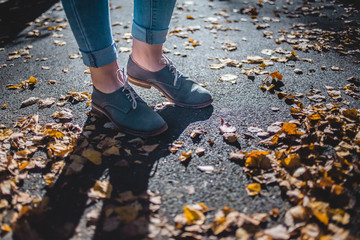 Women in cadet blue leather boots standing on yellow autumn birch leaves on asphalt. Her jeans are tucked up.