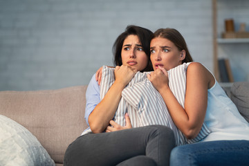 Two Scared Girls Hugging Pillows Sitting On Couch At Home