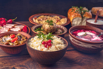 Ukrainian national cuisine - sauerkraut, donuts with garlic, canned mushrooms, borsch with sour cream, dumplings, baked potatoes, salted hot peppers in clay dishes on a dark wooden background. 
