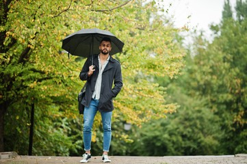 Fashionable tall arab beard man wear on black coat with umbrella and bag case posed at rain weather day.