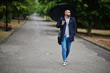 Fashionable tall arab beard man wear on black coat with umbrella and bag case posed at rain weather...