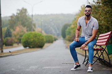 Obraz premium Fashionable tall arab beard man wear on shirt, jeans and sunglasses posed on park at bench.