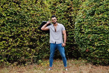 Fashionable tall arab beard man wear on shirt, jeans and sunglasses posed on park and hold bag case.
