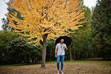 Fototapeta premium Fashionable tall arab beard man wear on shirt, jeans and sunglasses posed on autumn park with umbrella against yellow leaves tree.