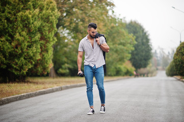 Fashionable tall arab beard man wear on shirt, jeans and sunglasses walking at park with umbrella and coat at hand.