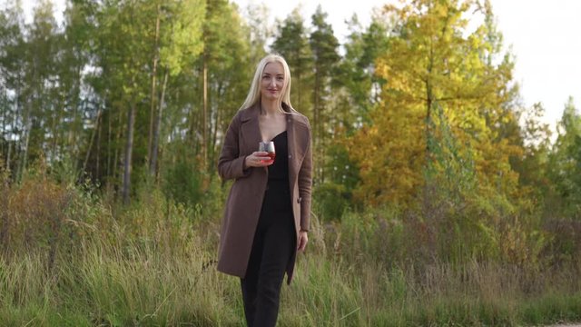 Long shot: Blonde woman drinking whisky cognac from a glass outside autumn park with golden and green forest in the background - Wearing a brown coat