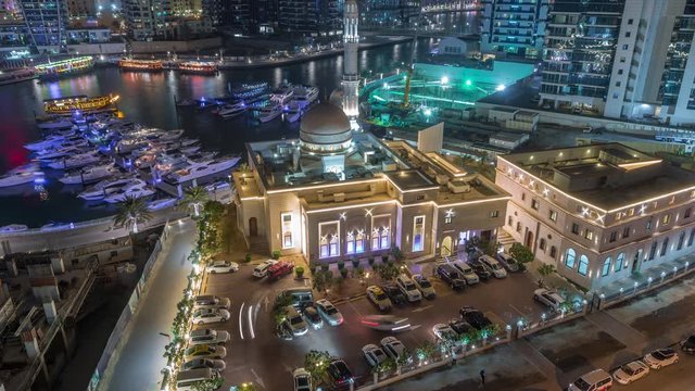 Yachts in Dubai Marina flanked by the Al Rahim Mosque with parking and residential towers and skyscrapers aerial night timelapse. Modern high rise skyline with boats and wooden dhows