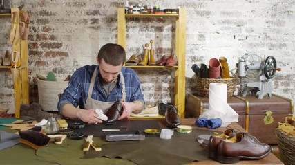 Tracking shot of professional male shoemaker in apron sitting at desk in workshop and polishing leather boot with cloth wrapped around fingers