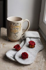 Grey knitted cozy socks on the windowsill, with a dry roses and a mug . A cozy home atmosphere.