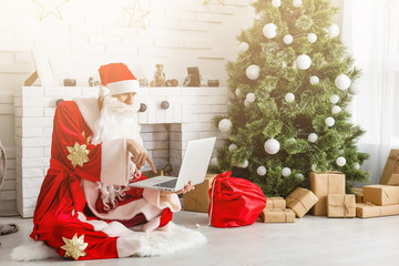 Fototapeta premium Modern Santa Claus. Cheerful Santa Claus working on laptop and smiling while sitting at his chair with fireplace and Christmas Tree in the background