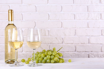 Obraz premium Vintage bottle of white wine without label, two glasses and bunch of grapes on wooden table, lofty white brick wall background. Expensive bottle of chardonnay concept. concept. Copy space.