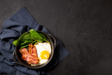 Fried egg with slices of bacon and spinach leaves in a black pan with handles on a black...