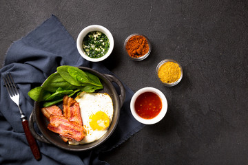 Fried egg with slices of bacon and spinach leaves in a black pan with handles on a black background. Classic English breakfast. Top view with copy space for your text