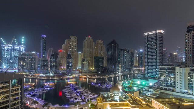 Yachts in Dubai Marina flanked by the Al Rahim Mosque and residential towers and skyscrapers aerial timelapse during all night with lights switching off. Modern high rise skyline with boats and wooden