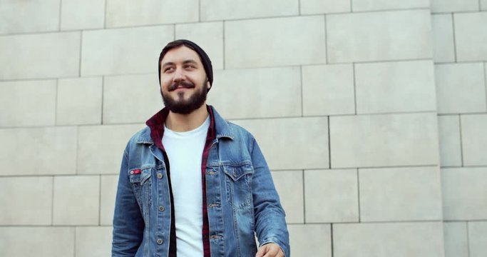 Portrait shot of young Caucasian handsome and stylish guy hipster smiling to the camera while standing at the wall outside, then cute happy girl coming from behind and jumping on his back cheerfully.
