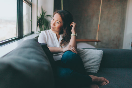 A Beautiful Asian Woman Sitting On The Couch While Looking Out The Window.