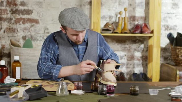 Professional shoemaker dyeing leather boot with brush while working at desk in his workshop