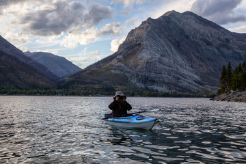 Adventurous Man Kayaking in Glacier Lake surrounded by the beautiful Canadian Rocky Mountains during a cloudy summer sunset. Taken in Upper Waterton Lake, Alberta, Canada.