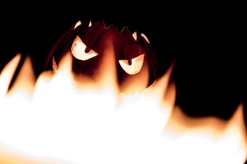 Spooky smiling halloween pumpkin in hot burning hell fire flames. The big helloween symbol has a mad face glowing eyes and also a glow in its mouth and teeth. Black orange nightmare of October 31st