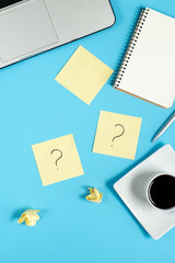 Question mark on sticky notes, notebook, cup coffee, laptop and pen on blue background. FAQ concept. Working with clients. Troubles in work. Flat lay, top view, copy space