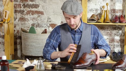 Tilt up of professional male shoemaker sitting at work desk in his studio, polishing leather boot with brush and then looking at camera and smiling