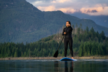 Adventurous Girl on a Paddle Board is paddling in a calm lake with mountains in the background during a colorful summer sunset. Taken in Stave Lake near Vancouver, BC, Canada.