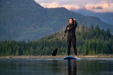 Adventurous Girl on a Paddle Board is paddling in a calm lake with mountains in the background during a colorful summer sunset. Taken in Stave Lake near Vancouver, BC, Canada.