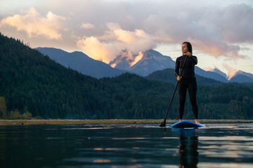 Naklejka premium Adventurous Girl on a Paddle Board is paddling in a calm lake with mountains in the background during a colorful summer sunset. Taken in Stave Lake near Vancouver, BC, Canada.