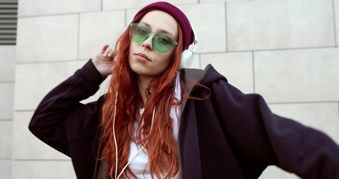 Cool stylish young Caucasian hipster girl with long red hair, sunglasses and headphones listening to the music and dancing. Outside.