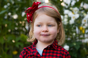 Medium horizontal portrait of pretty blue-eyed blond four-year old girl in red and black checkered shirt looking sideways with cheeky expression while standing in garden