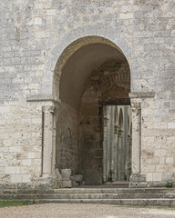 White stone wall with arched entryway, with broken sculpture, and columns visible beyond at the ruined 11th century Jumieges Abbey in Normandy France.