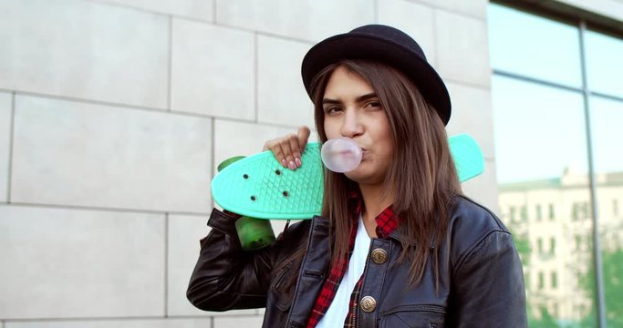 Portrait shot of the young stylish good looking Caucasian woman in hipster style and black hat holding her skateboard on the shoulder, looking straight to the camera and blowing bubble gum. Outdoor.