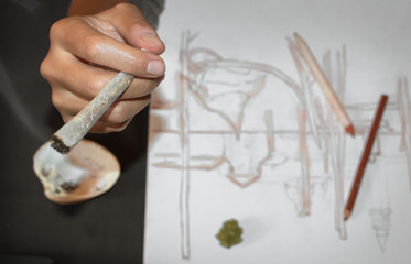 Smoking a joint of marijuana during a pause after drawing. Close up of hands and joint, blur effect. Background with pencils and sketches, copyspace.
