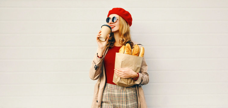 Portrait beautiful woman drinking coffee holding grocery shopping paper bag with long white bread baguette over blank gray wall background