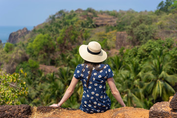 girl in a hat sits on the edge of a cliff overlooking the sea