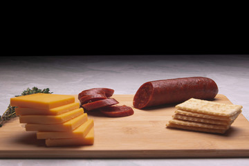 Sliced pepperoni on a bamboo cutting board with  cheddar cheese and crackers on a marble table.