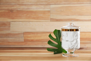 Decorative glass jar with cotton pads and monstera leaf on table against wooden background. Space for text