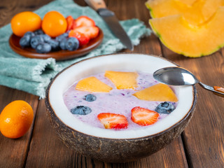 Close up view of blueberry smoothie in a coconut bowl with strawberries and melon and kumquat on a green towel with a teaspoon and knife. Soft focus.