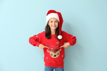 Little girl in Christmas sweater and Santa hat on color background