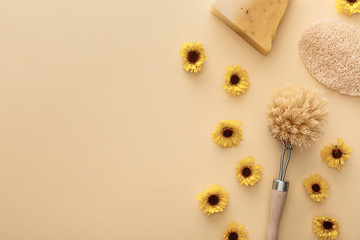top view of body brush, loofah and piece of soap on beige background with flowers and copy space