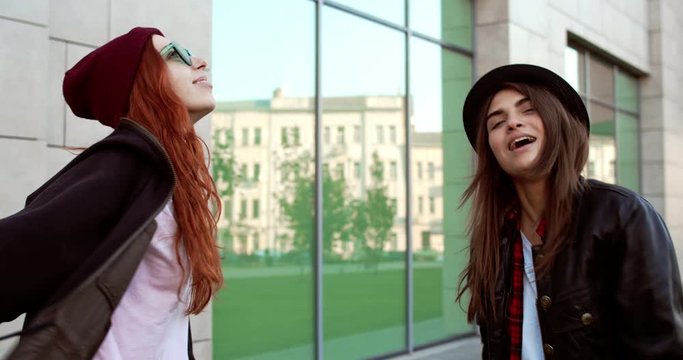 Pretty two Caucasian stylish hipster young femele friends hanging out together outside at some building, smiling and dancing.