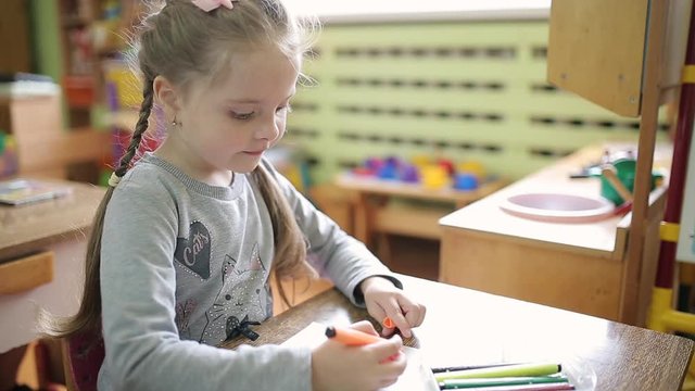 A girl draws a drawing on a white sheet while sitting at a table in kindergarten. 6 year old child engaged in pre-school education