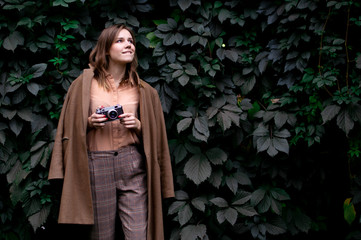 young girl photographer stands with a film camera near a wall of leaves in the forest, a woman...